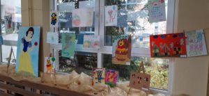 Zdjęcie przedstawiają prace plastyczne, które wpłynęły na konkurs. Galeria została zorganizowana w holu przedszkola. Prace zostały zawieszone na sznurku, na tle okna. Przedstawiają ulubione bajkowe postaci.