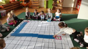 Dzieci siedzą wokół dużej planszy do kodowania, układają z niebieskich pól drogę od kotka do kwiatka.