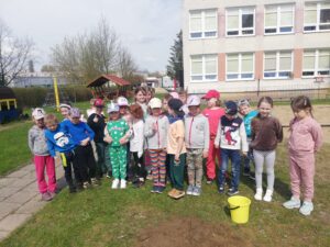Na zdjęciu znajduje się grupa dzieci zgromadzona w ogrodzie przedszkolnym wokół świeżo posadzonego drzewa