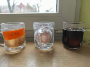 Na zdjęciu znajdują się trzy szklanki wypełnione wodą, coca colą i octem. W każdym kubku zatopione jest jajko. Szklanki stoją na stoliku