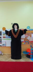 Na zdjęciu znajduje się postać Mikołaja Kopernika. Postać stoi na środku sali przedszkolnej. W tle znajdują się tablice  zawieszone na ścianie a na nich ilustracje 