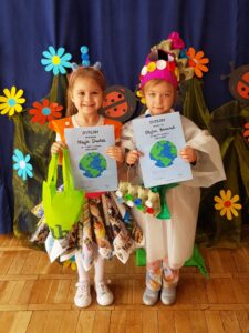Na zdjęciu dwie dziewczynki trzymające w rekach dyplomy (na niebieskim tle kula ziemska). Dzieci przebrane w stroje wykonane z materiałów nadających się do recyklingu.