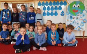 Na zdjęciu siedząca grupa dzieci ubranych na niebiesko. W tle papierowe kropelki wody oraz plakat o walorach wody.