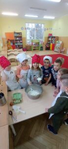 Na zdjęciu kilkoro dzieci przebranych w elementy stroju kucharza - czapki i fartuchy, stoją przy stole. Dziewczynka dodaje jogurt naturalny do miski z mąką i cukrem oraz jajkami.