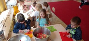 Na zdjęciu dzieci siedzące przy stoliku - dwie dziewczynki i jeden chłopiec. Na stole naczynia - miski metalowe, deski do krojenia oraz warzywa i składniki na sałatkę. Dzieci kroją warzywa.