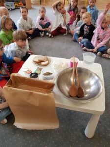Zdjęcie przedstawia grupę dzieci siedzących na dywanie, biały stolik na którym umieszczone są składki niezbędne do upieczenia chleba: mąka, woda, pestki dyni, miska, łyżki, blaszka, papier do pieczenia.
