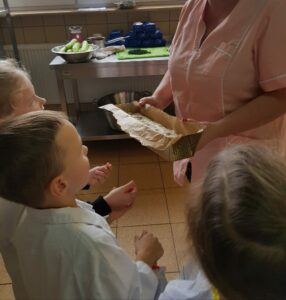 Na zdjęciu znajduje się grupa dzieci wraz z panią kucharką trzymającą w dłoniach blaszkę z przygotowanym ciastem na chleb.