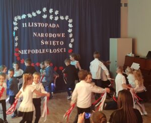 Na zdjęciu dziewczynki i chłopcy ubrani w stroje galowe, z biało-czerwonymi wstążeczkami w dłoniach tańczą w parach taniec patriotyczny.