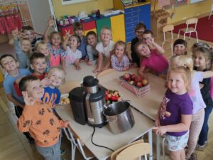 Grupka dzieci stojących wokół stołu, na którym ustawiona jest sokowirówka oraz owoce i warzywa. Dzieci są uśmiechnięte. Przygotowują się do zrobienia soków.