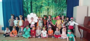 Na zdjęciu grupa dzieci przebranych za postacie zwierząt z lasu. Dzieci stoją i siedzą pod dekoracją lasu.
