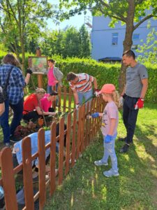 Dziewczynka w różowej czapce i koszulce trzyma pędzel i maluje na brązowo drewniany płotek. Mężczyzna ze szpadlem w ręku przekopuje ziemię. Dwie kucające kobiety sadzą rośliny. W tyle stoi kobieta w różowej bluzce i szarej marynarce.