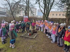 Dzieci z grupy V i VI stoją na trawniku na terenie przedszkola, w miejscu zasadzenia Dębu Katyńskiego i pomnika. Przedszkolaki zgromadzone są w półkolu, w centralnym punkcie zdjęcia znajduje się dąb katyński, kamień - pomnik oraz złożone dwa znicze - biały i czerwony