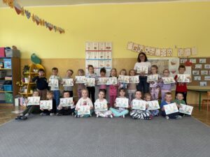 Na fotografii, na tle żółtej ściany i szarego dywanu znajdują się dzieci w dwóch rzędach: pierwszy rząd siedzi na dywanie, drugi rząd stoi. Wszystkie dzieci prezentują do aparatu kolorowanki o tematyce związanej z oszczędzaniem ciepła. Wśród dzieci w drugim rzędzie stoi pracowniczka olsztyńskiego MPEC, która przeprowadziła z przedszkolakami zajęcia.