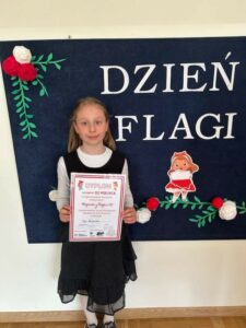 Zdjęcie przedstawia dziewczynkę 8-letnią na granatowym tle z napisem Dzień Flagi i trzymającą dyplom.