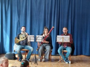 Na zdjęciu występują trzej muzycy grający na instrumentach - od lewej strony: mężczyzna grający na waltorni, kobieta grająca na fagocie i ostatni mężczyzna grający na klarnecie.