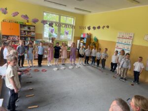 Na fotografii znajdują się ubrane odświętnie, stojące w półkolu dzieci. Przedszkolaki stoją na szarym dywanie, na którym rozłożone są instrumenty: klawesy, tamburyna, bębenki i janczary.