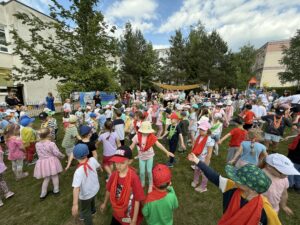 Zdjęcie wykonane na przedszkolnym placu zabaw. Na fotografii widać dużą grupę dzieci w kolorowych chustkach na szyi, symbolizujących poszczególne przedszkolne grupy. Dzieci biorą udział w zabawie ruchowej, prowadzonej przez tatę jednego z przedszkolaków. W tle stoją rodzice, w oddali widać również baner z danymi przedszkola i napisem "Festyn Rodzinny 2025"
