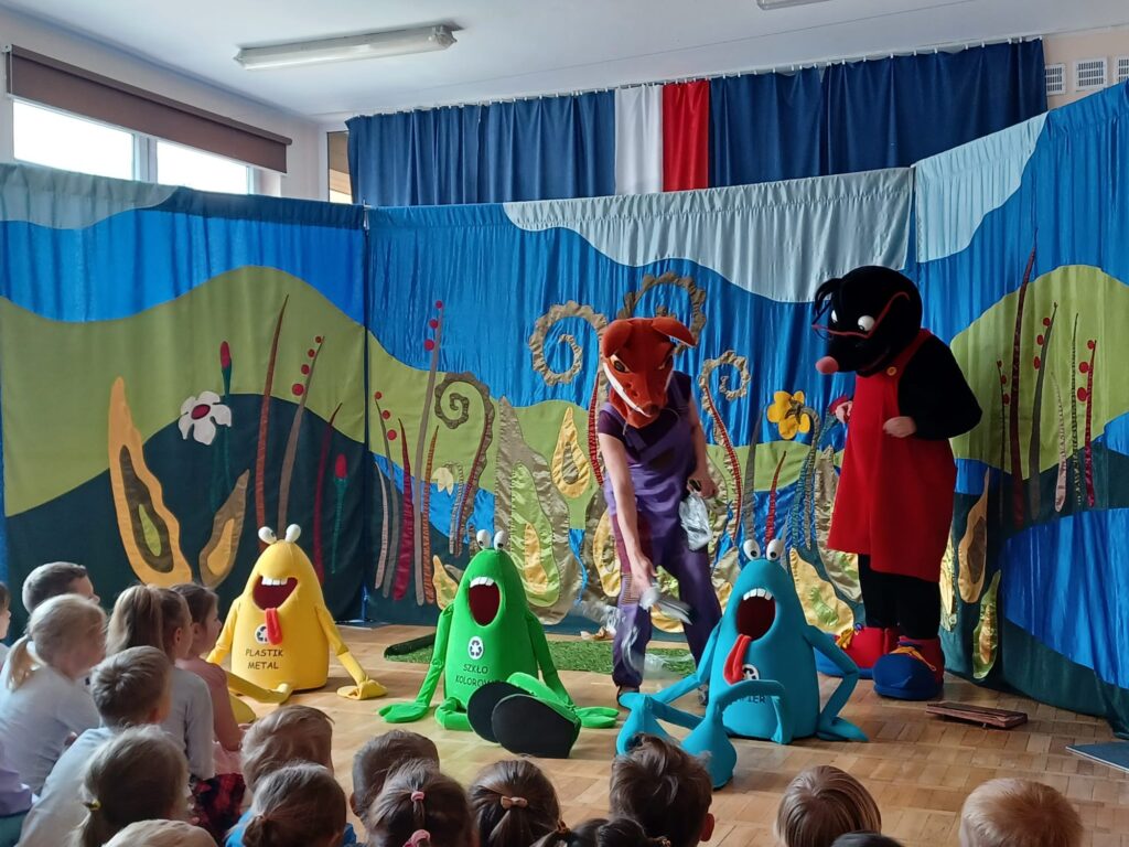 Zdjęcie przedstawia bohaterów przedstawienia- Krecika Ecika (po prawej stronie sceny) i Liska Kiteczkę (na środku sceny). Aktorzy stoją na tle makiety lasu, a przed nimi stoją tekstylne pojemniki na odpady w kolorze żółtym, zielonym i czerwonym. Lisek wkłada do niebieskiego pojemnika papier-do otworu w kształcie otwartej buzi.