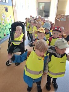 Dzieci stojące w parach, założone mają odblaskowe kamizelki. Dziewczynka na wózku w zielonej odblaskowej kamizelce. Niektóre dzieci mają na głowach czapki.