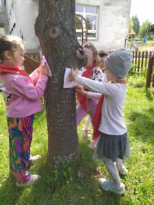 Na fotografii, w centralnej jej części widać pień drzewa, przy którym stoją cztery dziewczynki, kalkujące jego korę. W tle widoczny jest budynek przedszkola.