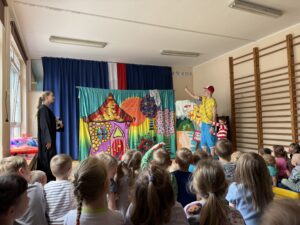 Na pierwszym planie znajdują się dzieci usadzone tyłem do fotografa. Na drugim planie widać tło teatralne przedstawiające kolorowy domek, drzewa oraz plażę i napis „zakaz kąpieli”. Na tym tle stoją aktorzy - po lewej stronie kadru stoi mężczyzna w żółtym T-shircie i niebieskich krótkich spodenkach, po prawej stronie kadru stoi kobieta w czarnej pelerynie