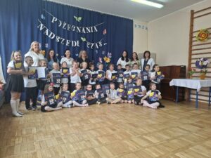 Grupa dzieci stojąca na tle dekoracji, ubrane na galowo, trzymają dyplomy i książki. Z tyłu dyrekcja , nauczycielki i pracownicy przedszkola.