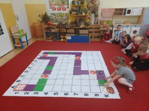 Zdjęcie przedstawia dziewczynkę układającą na macie do kodowania-rozłożonej na dywanie, kolorową płytkę w kwadratowym kształcie. Droga ta prowadzi od startu do koszyka z uwzględnieniem jabłek rozmieszczonych w dowolnych miejscach maty.