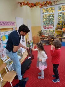 Zdjęcie przedstawia Tatę Kajetana stojącego po lewej stronie sali przedszkolnej i trzymającego w dłoni skrzypce. Po prawej stronie stoi dziewczynka grająca palcami na skrzypcach oraz chłopiec czekający na możliwość wydobycia dźwięku z instrumentu.