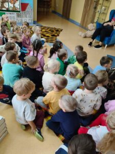 Na zdjęciu widać dzieci siedzące tyłem do fotografa w holu głównym przedszkola. Dzieci słuchają wierszy Wandy Chotomskiej.