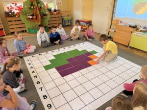 Na zdjęciu widać dużą białą matę do kodowania wokół której siedzą dzieci. Przedszkolaki układają na planszy kolorowe pola według kodu, by ułożyć sylwetę żołędzia. Na planszy kuca jeden z chłopców, który kładzie kolorowy kwadrat na planszy.