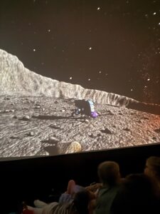 Na zdjęciu widać dzieci siedzące w namiocie sferycznym, na suficie namiotu wyświetla się animacja o kosmosie. Widać fragment skalistej planety i gwieździste niebo.