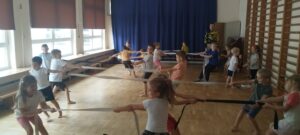 Na zdjęciu widać grupę dzieci w sali gimnastycznej podczas ćwiczeń. Dzieci w parach przeciągają pasy.