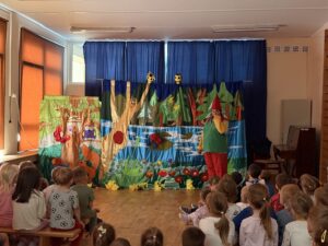 Na fotografii widać planszę przedstawiającą las, na tle której stoi aktorka przebrana za krasnala. Na pierwszym planie widać siedzące tyłem do fotografa dzieci, oglądające spektakl.