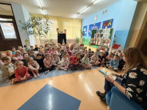 Na fotografii widać dzieci z grupy IV, V i VI, siedzące po turecku na kocach w holu głównym przedszkola. Dzieci słuchają nauczycielki siedzącej w niebieskim fotelu, czytającej utwory patronki przedszkola.