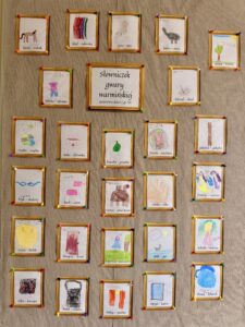 Fotografia przedstawia tablicę, na której znajdują się rysunki dzieci w złotych ramkach. Rysunki przedstawiają m.in. gęś, psa, królika, sukienkę itp. Tablica jest zatytułowana „słownik gwary warmińskiej autorstwa dzieci z gr. VI