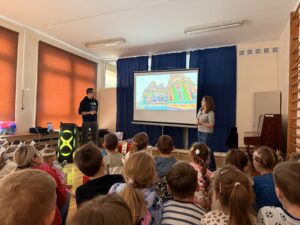 Na zdjęciu widać salę gimnastyczną, w której siedzą dzieci oglądające prezentację multimedialną wyświetlaną przez parę podróżników. Na tablicy wyświetlany jest slajd przedstawiający azjatycką świątynię o wielobarwnych ścianach.