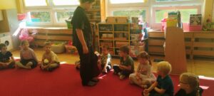 na zdjęciu widać dzieci siedzące w siadzie skrzyżnym w półkolu, podczas gdy starszy chłopiec pokazuje siedzącym przedszkolakom gitarę akustyczną.