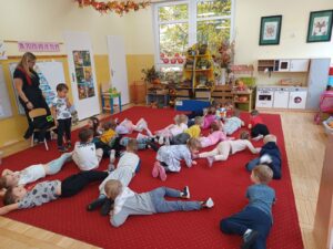 Zdjęcie przedstawia grupę przedszkolaków ćwiczących czołganie się pod okiem Mamy będącej członkiem Ochotniczej Straży Pożarnej.