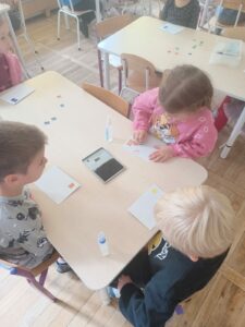 Zdjęcie przedstawia czworo dzieci siedzących przy stoliku. Przed dziećmi leżą koperty z naklejonymi kolorowymi znaczkami. Jedno z dzieci-dziewczynka, stempluje znaczek na kopercie.