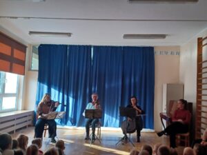 Zdjęcie przedstawia troje muzyków grających na skrzypcach, altówce i wiolonczeli. Po prawej stronie siedzi prowadząca koncert.