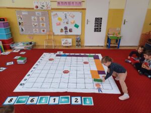 Choroba! Napiszę jeszcze raz, bo napisałam bez spacji.. Zdjęcie przedstawia matę do kodowania rozłożoną na dywanie. Dzieci stworzyły na macie ścieżkę prowadzącą od listonosza do dziecka (adresata). Dziewczynka po prawej stronie zdjęcia układa ostatnią kolorową płytkę.