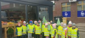 Na zdjęciu grupa dzieci stojąca przed osiedlowym warzywniakiem. Niektóre dzieci unoszą do góry torby z zakupionymi owocami i warzywami. Wszystkie dzieci ubrane są w kamizelki odblaskowe.