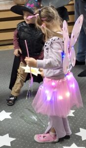 Na dywanie stoi chłopiec ubrany w czarną pelerynę z kapeluszem czarodzieja koloru czarnego oraz dziewczynka z różowymi skrzydłami wróżki i różową spódnicą z ledami. Na podłodze leżą białe gwiazdki z wróżbami.