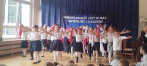 Na fotografii widać ubrane na galowo dzieci stojące w rozsypce. Dzieci stoją z rękami uniesionymi w górze, w doniach trzymają szyfonowe chusteczki w kolorze białym i czerwonym. Jest to kadr z tańca patriotycznego.