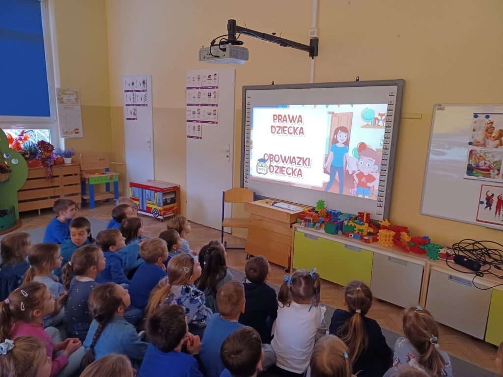 Zdjęcie przedstawia dzieci siedzące przed tablicą interaktywną. Wyświetlany jest film edukacyjny dotyczący praw i obowiązków dzieci.
