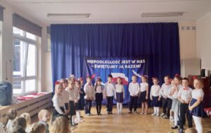 Na zdjęciu widać dzieci w strojach galowych ustawione w półkolu. Zdjęcie zostało zrobione podczas śpiewania hymnu narodowego. W tle znajduje się granatowa kotara z napisem "Niepodległość jest w nas, świętujmy ją razem"