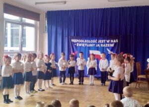 Na fotografii widać ubrane na galowo dzieci stojące w półkolu. Dzieci trzymają w dłoniach białe róże, śpiewają pieśń "Rozkwitały pąki białych róż". W tle - granatowa kotara z napisem "niepodległość jest w nas, świętujmy ją razem"