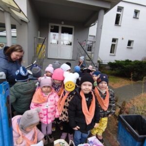 Na zdjęciu stojące dzieci w gromadce przed szarym budynkiem stowarzyszenia "Niebieski parasol". Przed dziećmi ustawione są cztery duże torby prezentowe wypełnione herbatami i słodyczami w opakowaniach kartonowych.