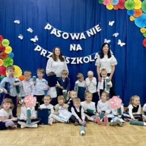 Na zdjęciu dwie nauczycielki oraz grupa dzieci siedzących i stojących w gromadce, pozują do pamiątkowego zdjęcia. w tle dekoracja na granatowym tle: powieszone litery napisu "pasowanie na przedszkolaka" oraz przyczepione białe motylki i w dwóch rogach kolorowe rozety