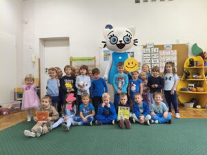 na zdjęciu widać grupkę dzieci ustawionych w dwóch rzędach. Za przedszkolakami stoi duża maskotka Kici Koci - białego kota z niebieskimi oczami i w niebieskiej sukience.